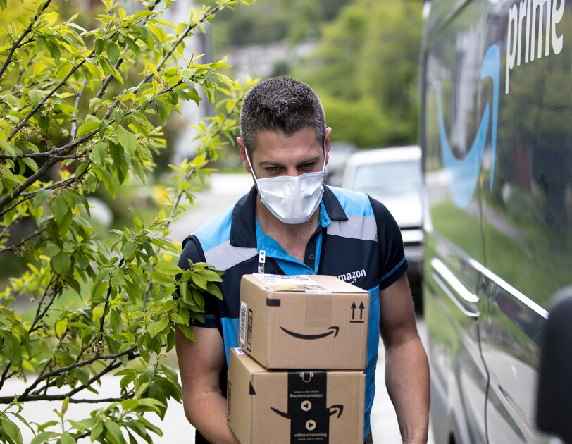 A delivery driver wearing a face mask walks alongside an Amazon Prime delivery van, carrying packages.