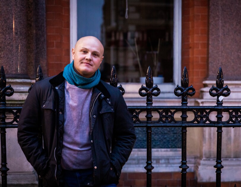 Man wearing black jacket with hands in pockets stood against some black railngs