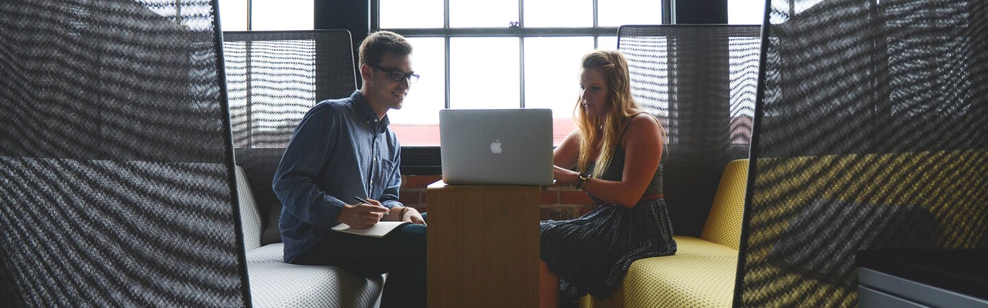 Man taking notes as his female colleague works on a laptop, in a casual work space.
