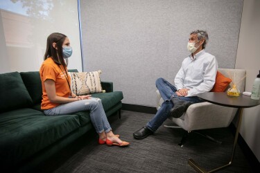 A man and a woman sit in a clinic setting, wearing masks, while speaking.