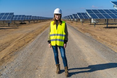 Eine dunkelhaarige Frau mit gelber Warnweste steht vor einem Solarpark. Sie lächelt in die Kamera. Hinter ihr sieht man die Solarpanels.