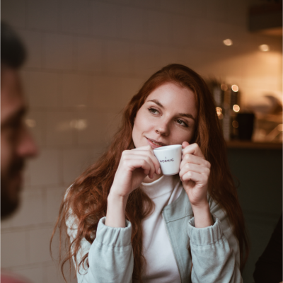 Eine rothaarige Frau sitzt an einem Tisch und hat eine weiße Espressotasse in den Händen. Sie trägt eine hellblaue Jacke und ein weißes T-shirt. Es sind verschwommen weitere Personen zu sehen, die Kaffeebecker in den Händen halten.