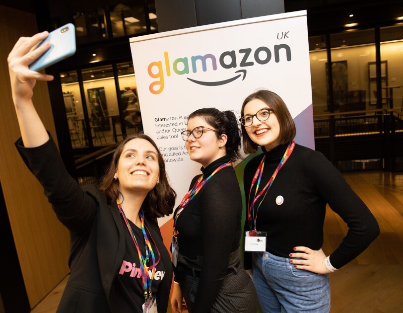 Two Amazon employees taking a selfie with a representative from PinkNews at the 2020 British LGBT+ Awards. They are standing in front of a glamazon banner.