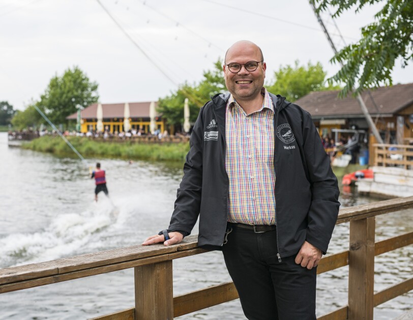 Ein man im karierten Hemd steht vor der Wasserski-Anlage und lächelt in die Kamera.