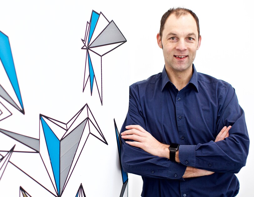 Ralf Herbrich, co-head of Amazon's Core Artificial Intelligence (AI) group. He wears a navy button down and leans against a white background with blue and gray illustrations.