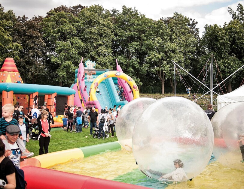 Verschiedene Spielstationen auf dem Spielfest: Man sieht Besucher und riesige Ballon, die auf einem aufblasbaren Schwimmbecken treiben, Im Balloninneren sind Kinder.