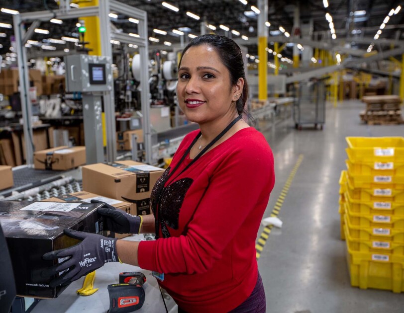 An Amazon associate holds a product near a conveyor belt. Tools sit on the countertop near her. She wears a red long-sleeve tee shirt and work gloves as she works at a Columbus, Ohio fulfillment center, CMH1