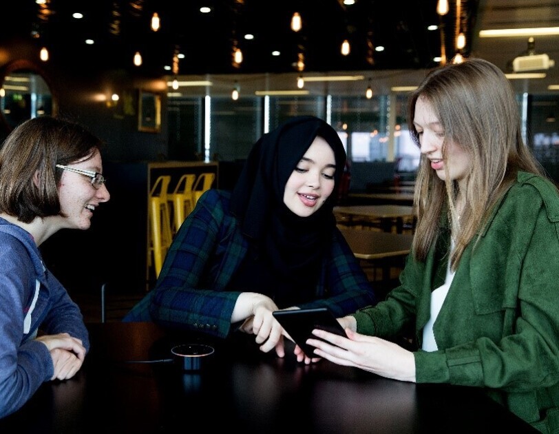 Three female students sat at a table smiling, talking and looking at a Kindle