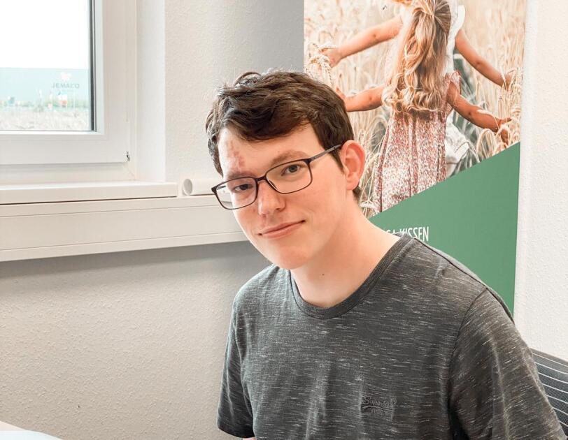 Ein Mann mit grauem T-shirt sitzt vor einem Computer und lächelt in die Kamera.