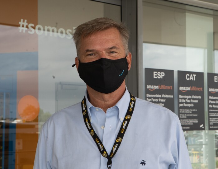 Fred Pattje, Director de Amazon Customer Fulfillment en Francia, Italia y España. De fondo el exterior de un centro logístico con unos carteles negros y letra blanca en español, catalán e inglés. También un dispensador de gel y una puerta de color azul. El protagonista va con una mascarilla corporativa de Amazon. Una camisa de color azul. Está moreno y el pelo es castaño y blanco con los ojos marrones.