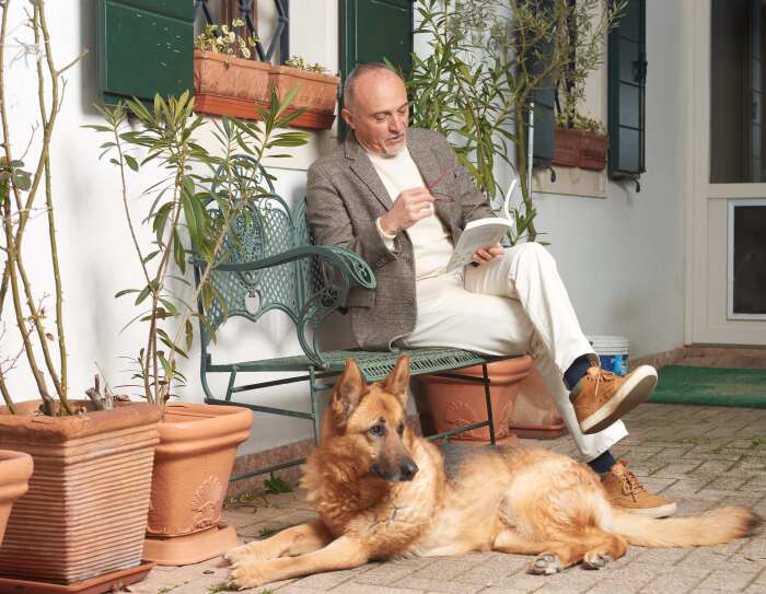 Lo scrittore Federico Maria Rivalta legge un libro sul portico di casa con un pastore tedesco ai suoi piedi.