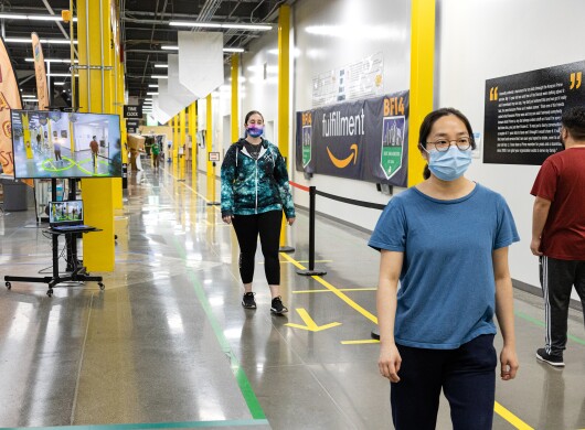 Amazon associates wearing face masks walk along a corridor of a fulfillment center. Behind them, a screen shows visual social distance assistance.