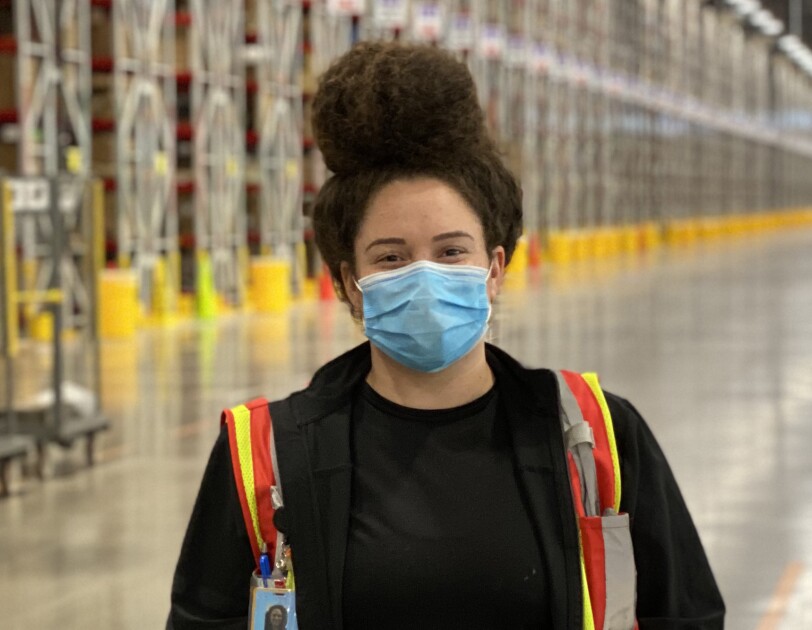 A woman wearing a safety vest and a mask stands in a warehouse space.