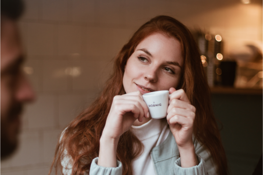 Eine rothaarige Frau sitzt an einem Tisch und hat eine weiße Espressotasse in den Händen. Sie trägt eine hellblaue Jacke und ein weißes T-shirt. Es sind verschwommen weitere Personen zu sehen, die Kaffeebecker in den Händen halten.