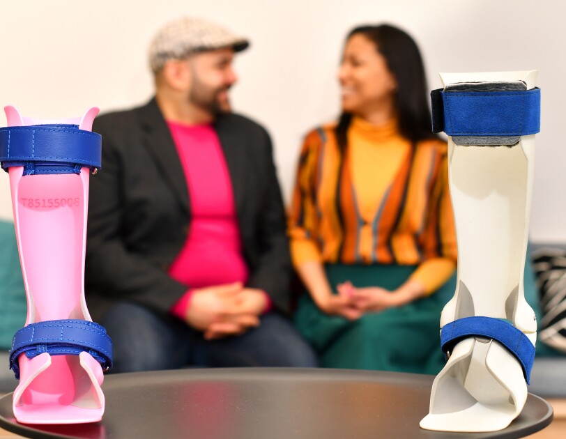 In the forefront, two ankle orthoses for children produced by Andiamo, a smaller one in bright pink plastic and a bigger one in white plastic with blue straps. In the background, Samiya and Naveed Parvez smile at each other.