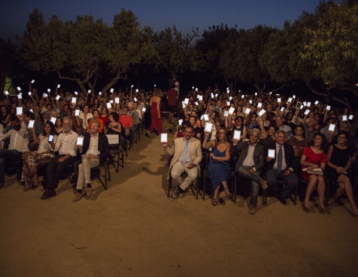 Platea di persone sedute nella Valle dei Templi di Agrigento durante un reading di Luigi Pirandello in occasione dei 150 della sua nascita. Gli spettatori reggono con una mano un Kindle PaperWhite, con lo schermo rivolto verso la camera. Sullo sfondo, degli alberi.