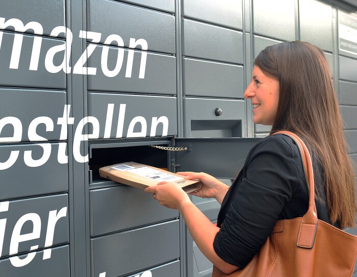Der Bildausschnitt zeigt graue Paketschließfächer und den weißen Schriftzug "Amazon bestellen hier ...". Rechts ist eine junge Frau mit langen braunen Haaren zu sehen, die ein längliches, flaches Amazon Paket aus dem Schließfach entnimmt.