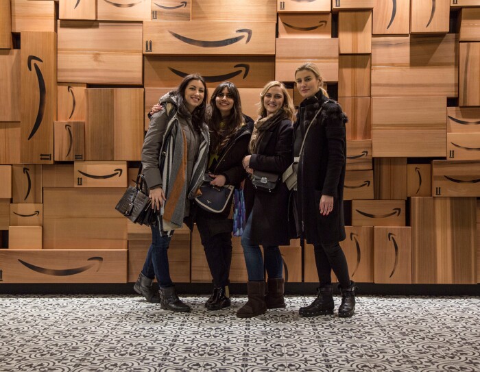 Quattro ragazze sorridono di fronte a un muro di scatole Amazon. Sono Monika, Urte, Francesca e Ankita, vincitrici dell'Amazon Design Award, quattro studentesse del Master in Interior Design dello IED Milano hanno vinto gli Amazon Design Awards, una competizione in cui si chiedeva ai corsisti di ripensare i depositi di smistamento Amazon con uno sguardo alla praticità e al comfort dei dipendenti.