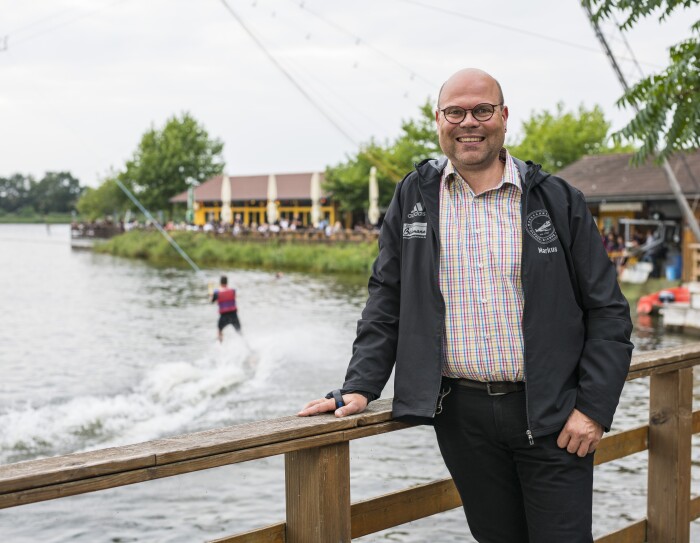 Ein man im karierten Hemd steht vor der Wasserski-Anlage und lächelt in die Kamera.