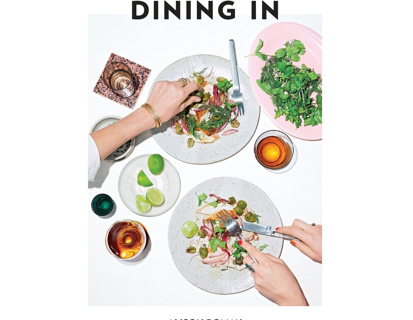 cover of a cookbook, called "Dining In" shows a woman with fork and knife poised to pick up items from her dinner plate, while another arm reaches across the table to grab an olive. drinks, lime slices and other accoutrements are tastefully organized.