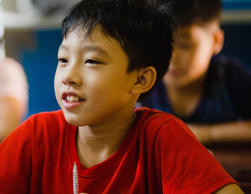 Ein asiatischer Junge im roten T-Shirt sitzt in der Schulbank. Im Hintergrund sieht man weitere Schulkinder, die am Unterricht teilnehmen.