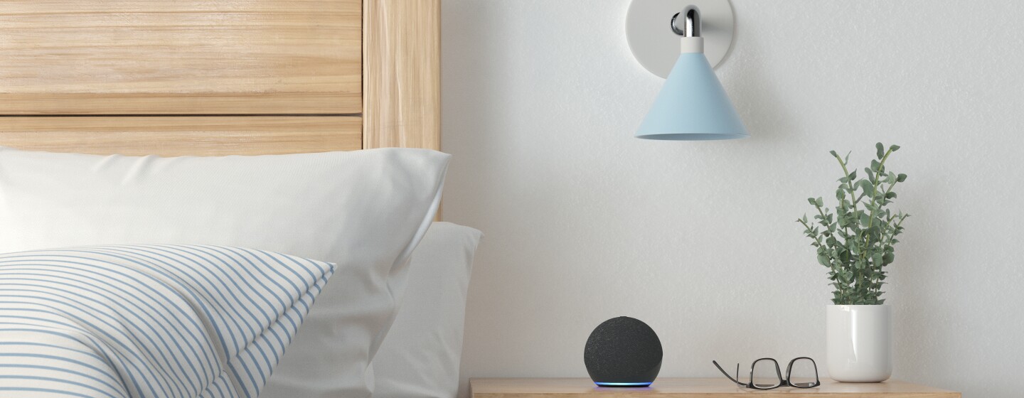 Una habitación con una cama y una mesita de noche. Los muebles son de madera, estilo pino. El cabecero es alto y la mesita de noche tiene dos cajones. En la cama dos cojines blancos y uno de rayas azules y blancas. Hay un plumón también con el mismo estampado. En la mesita de noche hay un cajón abierto con libros y otro cerrado. Encima de la mesita un Echo Dot de Amazon con la luz azul encenddia, una gafas, una planta verde y con un jarrón blanco. Y una lámpara de pared de color azul.