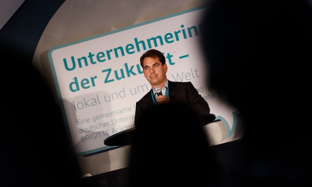 Nicholas Denissen steht auf der Bühne am Rednerpult. Er hält eine Laudation. Er ist aus dem Publikum heraus fotografiert worden. Im Vordergrund sind unscharfe Köpfe des Publikums zu erkennen.