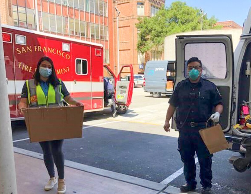 A woman wearing a safety vest and a protective mask carries a box into a fire station.