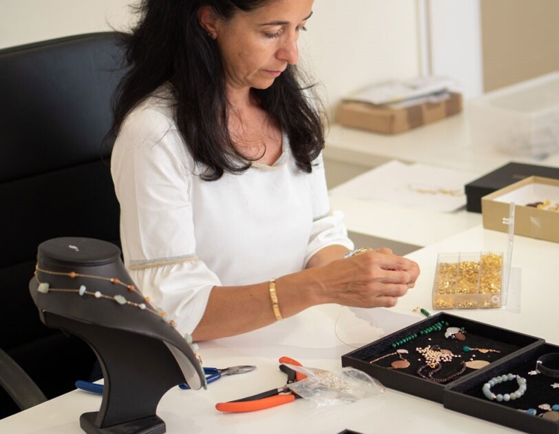 Maria A. Marinas sitzt an ihrem Schreibtisch und kreiert neue Schmuckstücke. Ihr Arbeitsplatz ist voll mit Perlen, Steinen, Drähten, Zangen und weiteren Materialien.