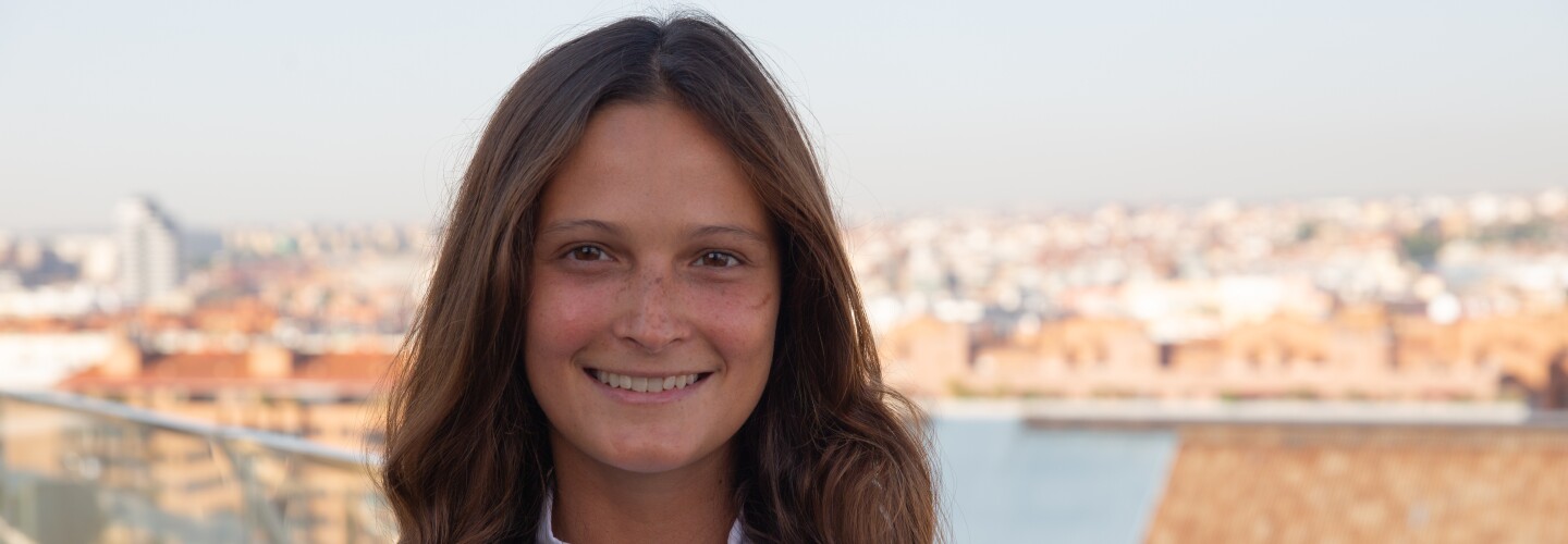 En la terraza de las oficinas logísticas de Amazon en Madrid, de fondo el skyline. En primer plano una mujer caminando decidida hacia la derecha de la imagen. Lleva el pelo suelto hasta el hombro y es de color castaño. Una camisa blanca y lleva la tarjeta de paso con un collar de color naranja. 