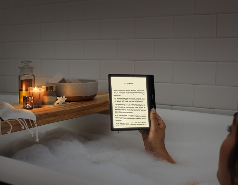A woman relaxes in a bubble bath while reading a Kindle Oasis in candle lighting.
