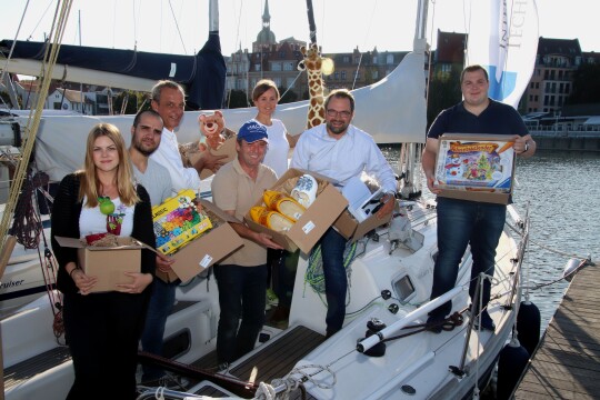 Eine Gruppe Menschen steht auf einem kleinen Segelboot. JEder hält einen Karton mit Spielzeug und Kuscheltieren in der Hand.