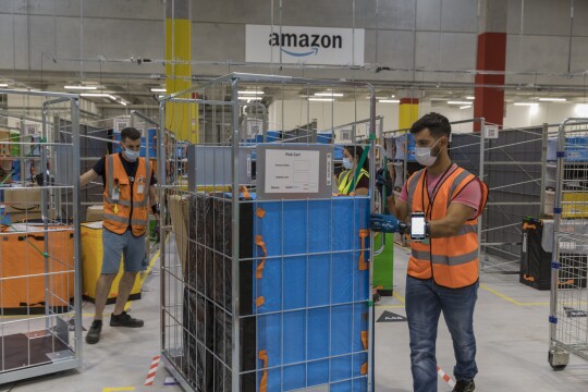 Un gruppo di dipendenti Amazon sposta dei carrelli contenenti dei pacchi. In alto sullo sfondo il logo di Amazon