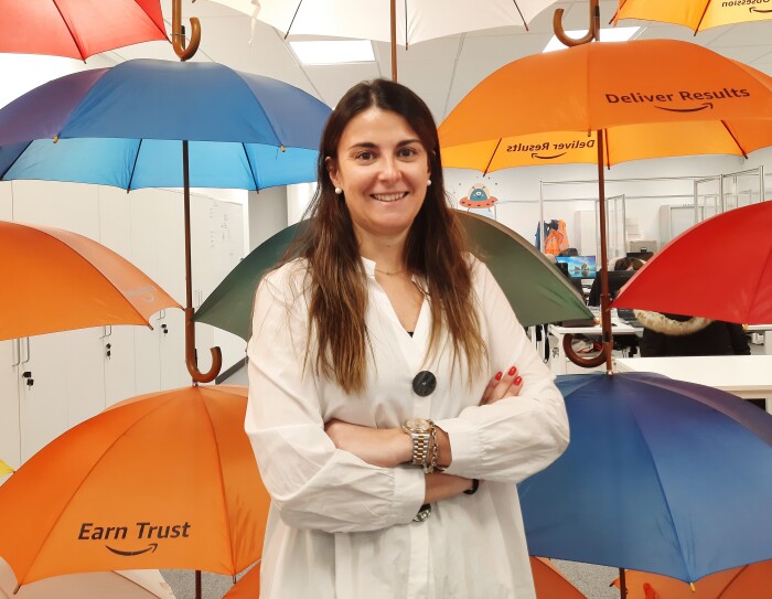 Raquel Mur. Va con una camisa blanca. Los brazos cruzados y mirando a cámara. Lleva las uñas rojas, unas pulseras en la mano izquierda. Tiene el pelo largo, liso y castañol. Mira a cámara sonriendo. El fondo es una oficina con paraguas que cuelgan del techo.