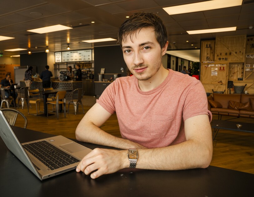 Jake Peters, 23, an Infrastructure Technician apprentice sitting in the UK Amazon corporate office cafeteria behind a laptop.