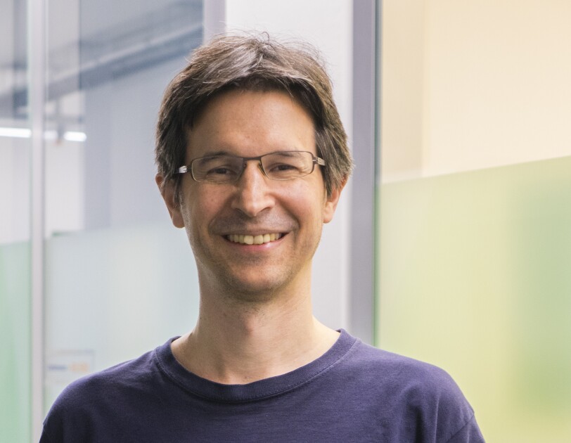 Ein Mann mit braunen Haaren, Brille und einem blauen T-shirt lacht in die Kamera.