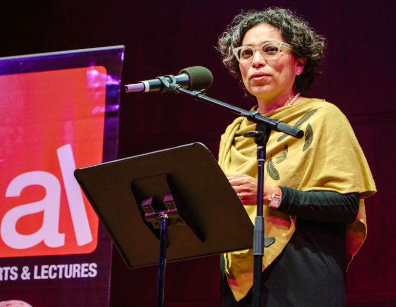 A woman standing at a lecture, in front of a portable podium and microphone. She appears to be speaking to a group, on a stage. In the background, Seattle Arts & Lectures logo is visible.