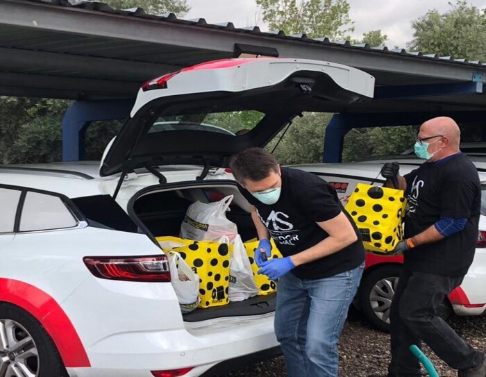 Manos de ayuda social reparte productos de primera necesidad gracias a la ayuda también de Securitas direct. Aparece en primer plano un coche de Securitas direct con dos de sus conductores que llevan una camiseta de Manos de ayuda socials de color negro. Los dos hombres de mediana edad llevan mascarilla y están colocando bolsas de color amarillo con lunares negro en el maletero de un coche. Están en un aparcamiento abierto.