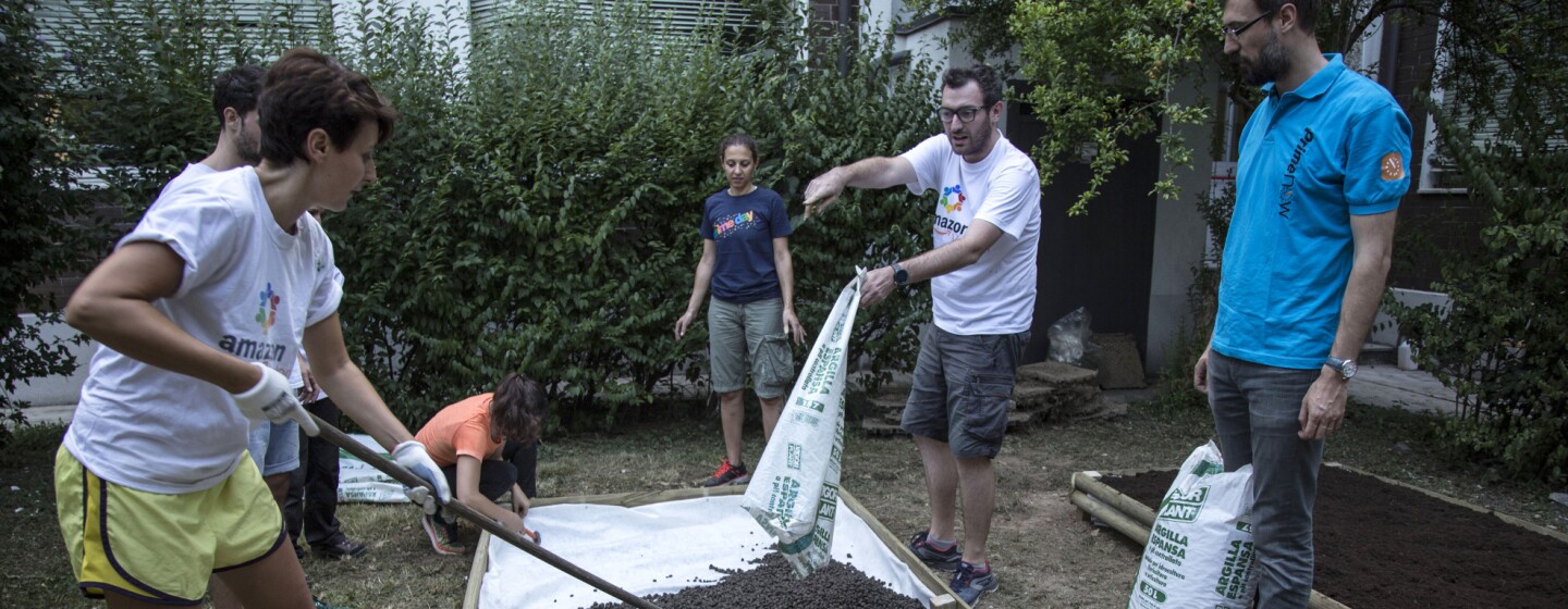 A scuola di verde: sei volontari Amazon all'opera nel giardino della scuola Media Leonardo da Vinci di Affori, nella periferia di Milano. Una donna in primo piano regge una pala con cui stende della terra, versata da un sacco da un uomo, in piedi di fronte alla donna. Gli altri 4 volontari osservano la scena.