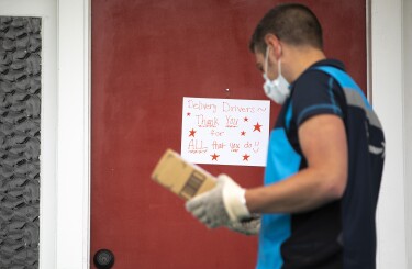 Amazon delivery driver stands in front of a door, to deliver a package. On the door is a sign that says "delivery drivers, thank you for ALL that you do."