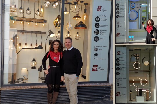Ein Mann und eine Frau stehen vor einem Schaufenster, in dem viele unterschiedliche Lampen zu sehen sind. Beide lächeln in die Kamera.