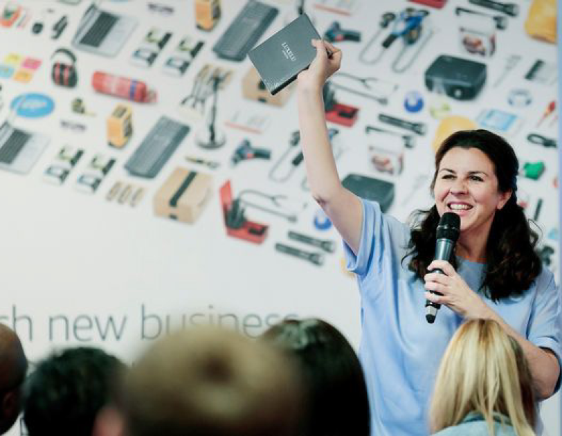 Woman with brown hair holding a mic and talking to an audience