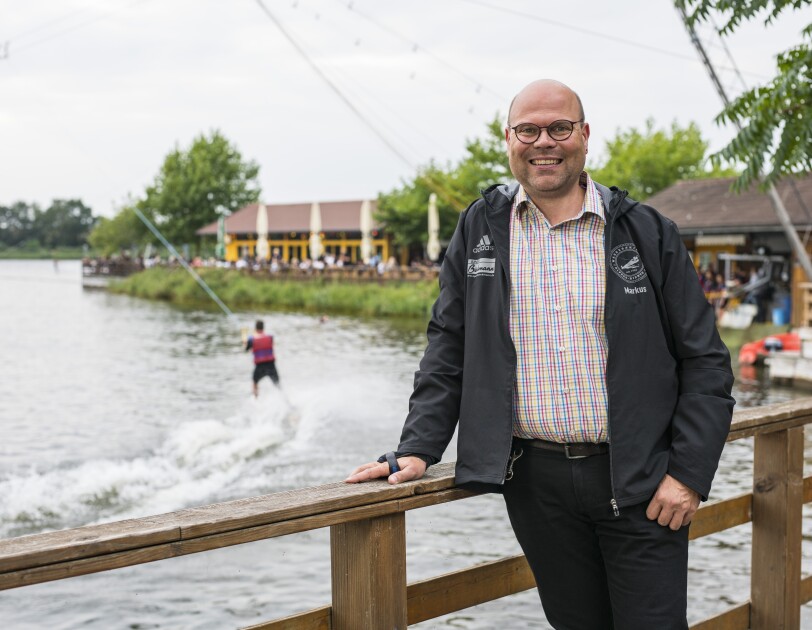 Ein man im karierten Hemd steht vor der Wasserski-Anlage und lächelt in die Kamera.