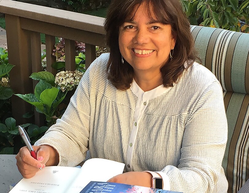 Amazon author Barbara Freethy sits on a deck in the sunshine behind a stack of her books. She is autographing one book.
