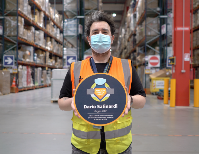 Uomo ripreso a mezzo busto in un magazzino Amazon. Indossa un giubbino catarinfrangente arancione e giallo, tiene tra le due mani e mostra alla camera un cartellone tondo con la scritta 'Dario Salinardi 2021' ed un logo che recita 'Amazon Safety Hero'. Indossa una mascherina chirurgica sul volto, ha i capelli neri con un ciuffo bianco. Alle sue spalle si intravedono gli scaffali di un magazzino Amazon.