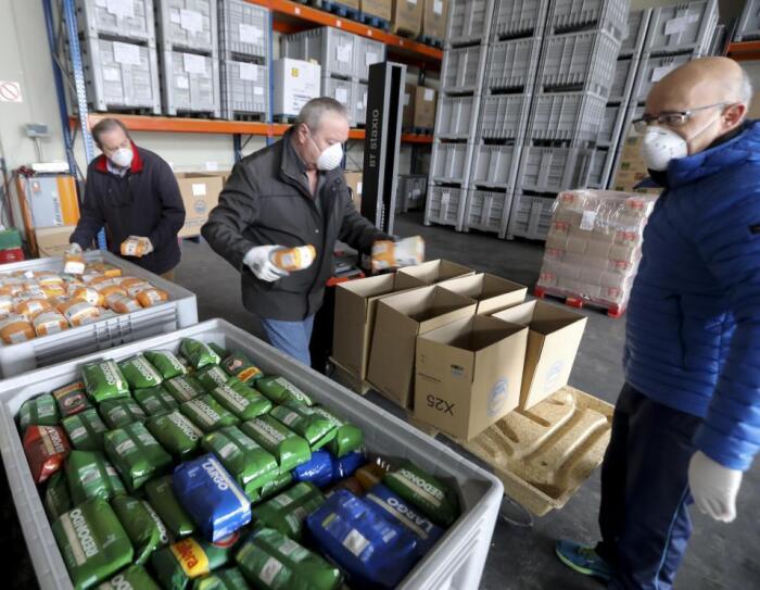 Banco de alimentos. En la parte inferior de la foto, dos cubetas grandes con paquetes de arroz y azúcar. Tres personas de mediana edad, con mascarilla, están con paquetes en las manos colocándolos en cajas de cartón. De fondo, estanterías llenas de palés de color gris.