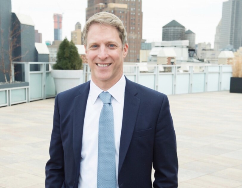 A man in a navy suit, white shirt, and slate blue tie stands outdoors in a city setting.