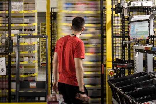 Un dipendente Amazon ripreso di spalle al centro dell'immagine osserva gli scaffali mobili del magazzino.