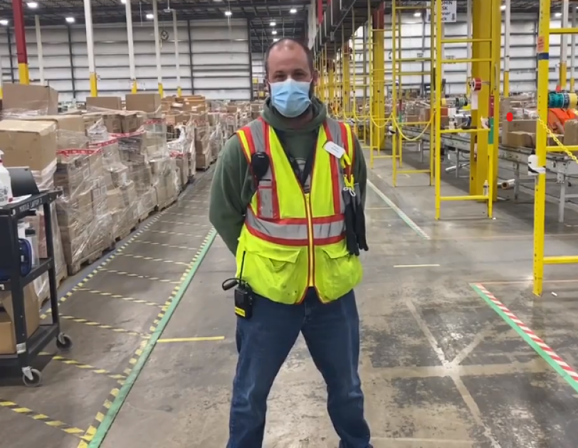 Russel Whitfield, an associate at Amazon, stands in an Amazon warehouse smiling for a photo. He is wearing a face mask and other Amazon warehouse safety gear.