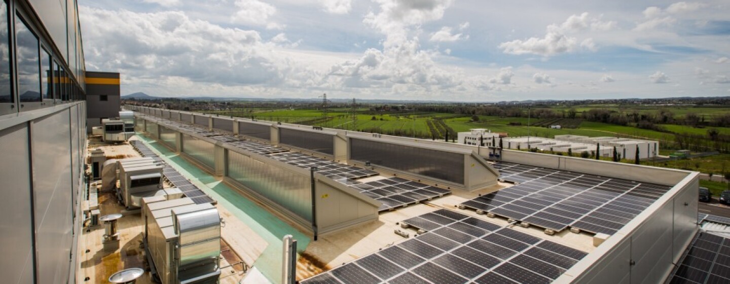 En primer plano aparece el tejado de un centro logístico con placas solares. Al fondo un paisaje de campos verdes y el cielo azul con nubes blancas.